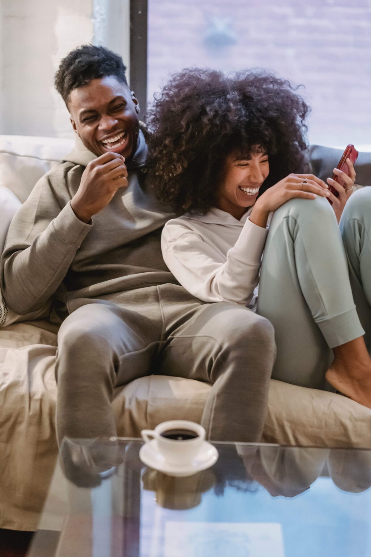 A couple sits closely on a couch, laughing together. One holds a smartphone while the other leans in. Both wear casual clothes, and a cup of coffee is on the glass table in front of them.