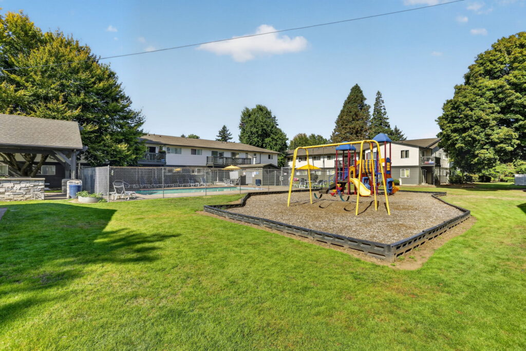 Playground with climbing structure and swings on mulch, surrounded by grass. Nearby are apartment buildings, trees, a covered area, and a fenced swimming pool under a blue sky.