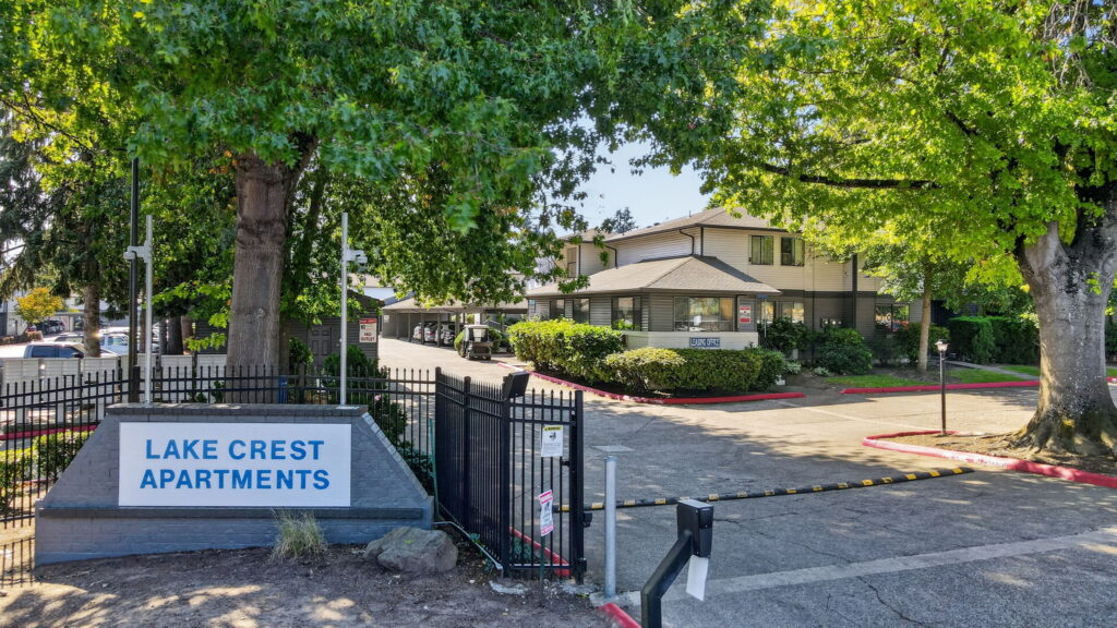 Entrance to Lake Crest Apartments, featuring a sign, gated entry, large green trees, and a two-story beige building in the background under a bright blue sky.
