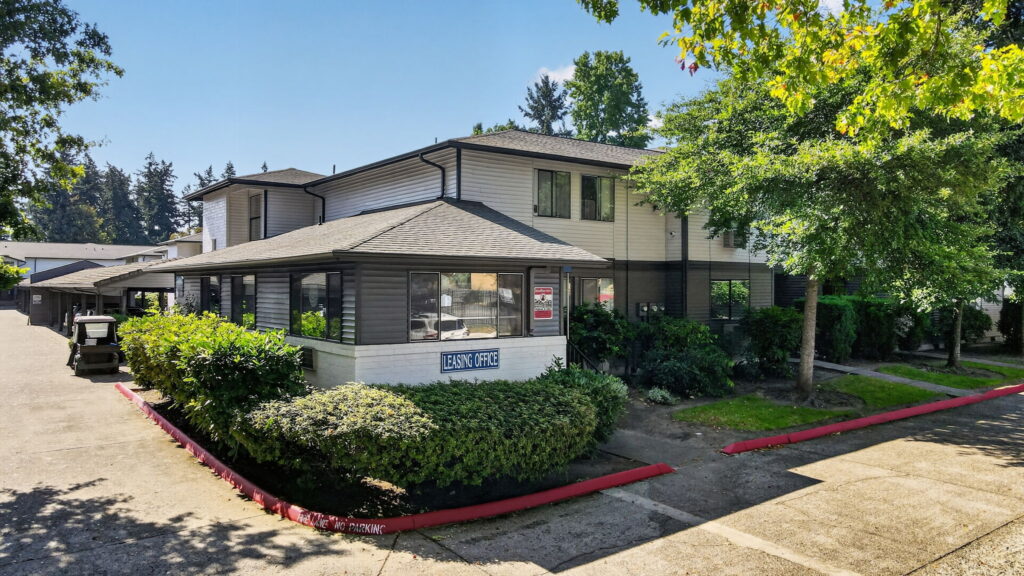 Two-story building with a sign reading Leasing Office in front, surrounded by green bushes and trees, under a clear blue sky.