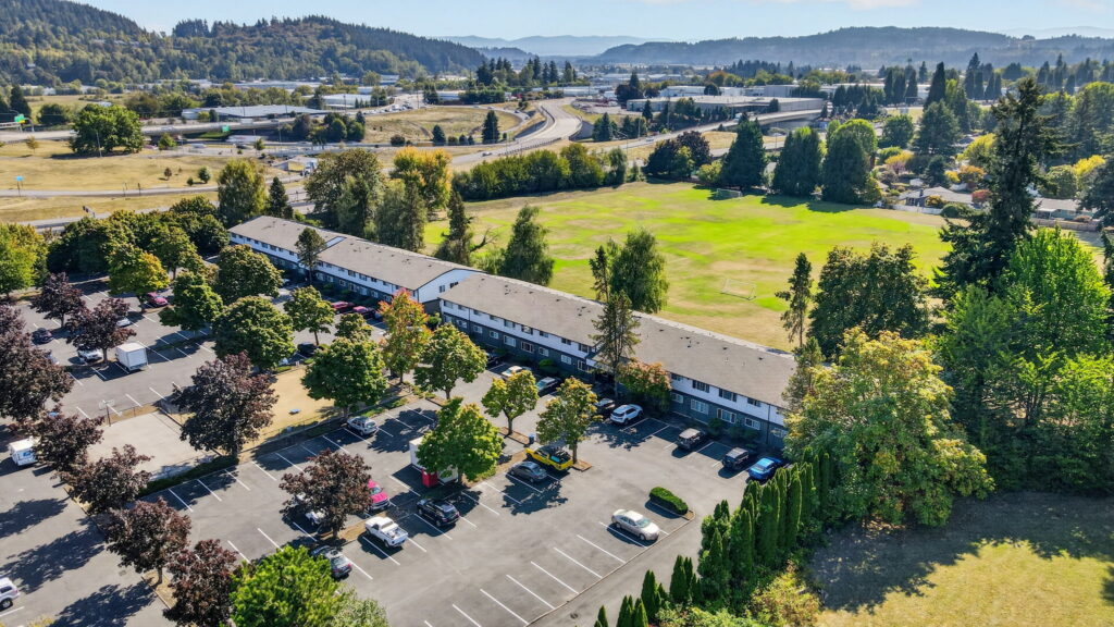 Aerial view of an apartment complex with cars parked, a large grassy field, trees, and a highway in the background, surrounded by green hills under a clear sky.