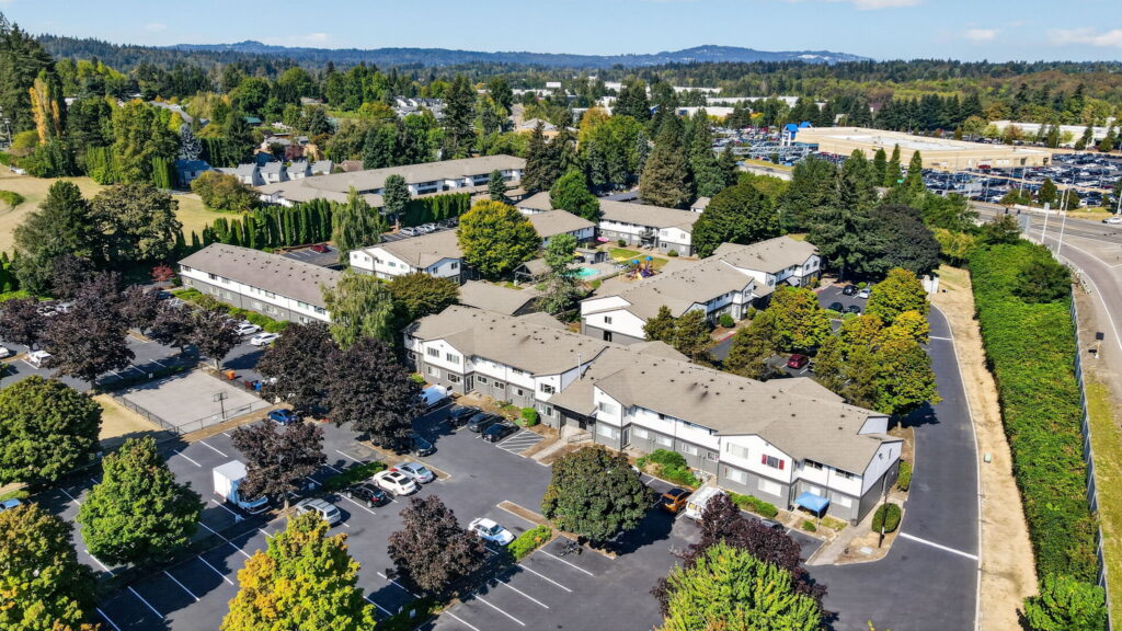 Aerial view of a suburban apartment complex surrounded by trees, parking lots with cars, and nearby roads, with a mix of residential and commercial buildings in the background.
