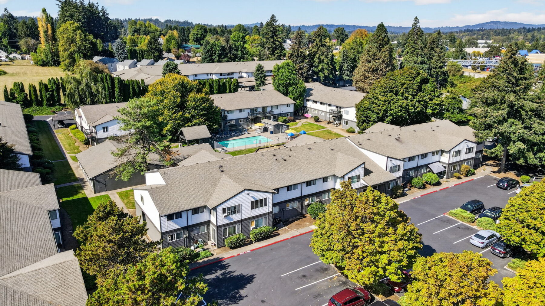 Aerial view of an apartment complex with multiple buildings, a central swimming pool, green lawns, trees, and parked cars in a lot on a sunny day.