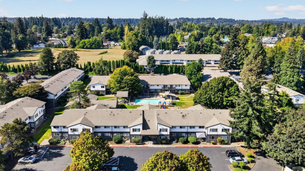 Aerial view of an apartment complex surrounded by trees, with a swimming pool and lounge chairs in the central courtyard on a sunny day.