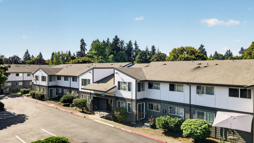 Two-story apartment building with gray roof, white and dark gray exterior, surrounded by trees and bushes, under a clear blue sky.