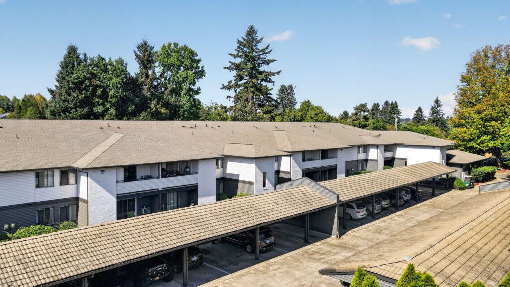 Aerial view of a two-story apartment complex with beige roofs, white and gray exterior, covered parking spaces, and trees in the background under a clear blue sky.